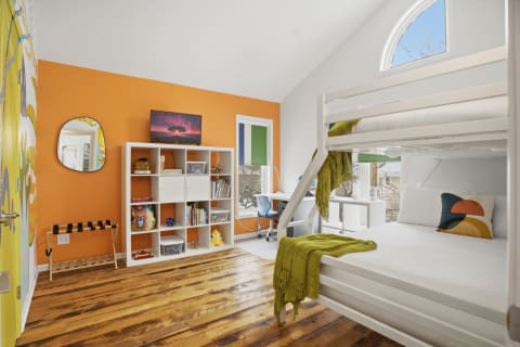 A bright bunk room with orange walls, a white bunk bed, colorful decor, and a workspace.