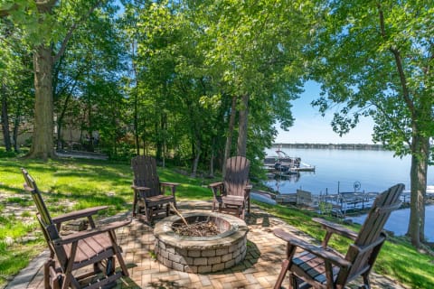 Adirondack chairs surround a fire pit next to a calm lake.