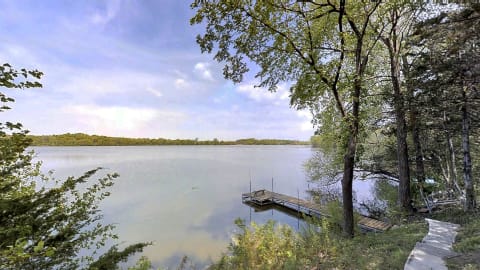 A tranquil lake scene with a wooden dock, surrounded by green trees under a cloudy sky.