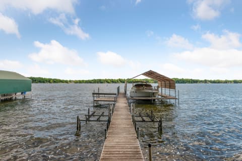 A wooden dock leading to a covered boat slip on a lake surrounded by green trees and a blue sky.