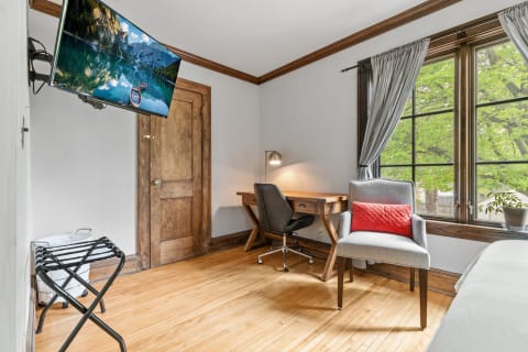 Interior of a room with a wooden desk, chair, television on the wall, and a window with greenery outside.