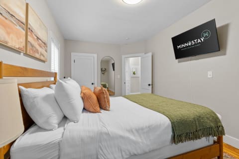 Cozy bedroom with white bedding, decorative pillows, green throw, and a wall-mounted TV.