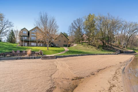 Lakeside property featuring a large house, sandy beach, and green landscape.