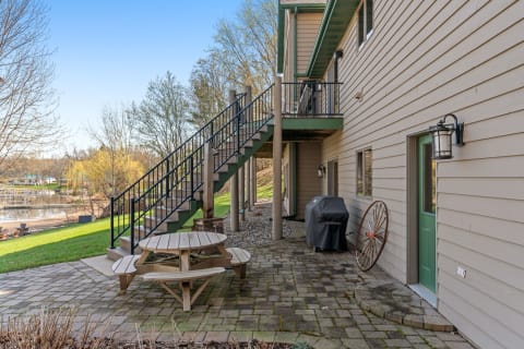 A patio area with a picnic table, grill, and steps leading to a balcony adjacent to a house, with a view of water and trees in the background.