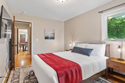 A cozy bedroom featuring a queen-sized bed with a red throw blanket, wooden nightstands, and a window with views of green trees.