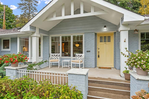 Bungalow-style home with a light blue exterior, yellow door, and a front porch adorned with white chairs and flowers.