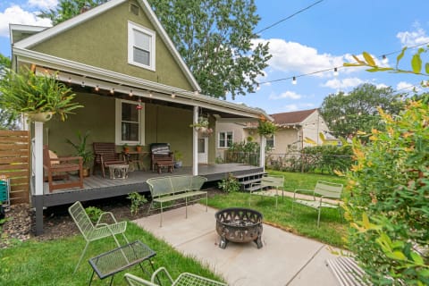 Backyard of a green house featuring a porch with chairs and a fire pit.