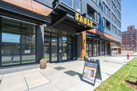 Modern apartment building entrance featuring the SABLE sign and sleek dark exterior design, with planters and a sales center sign.