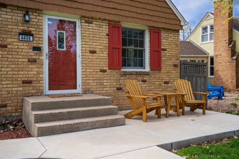 Charming brick house entrance with red door and wooden chairs.
