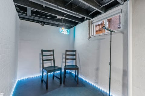 A small music room featuring two black chairs, a microphone, and a neon sign that reads 'Live Music'.