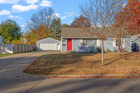 A single-story light blue house with a red door and garage, surrounded by trees in autumn.