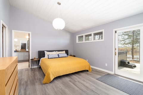 A contemporary bedroom featuring a queen bed with a yellow quilt, a dresser, and large glass doors leading to a view of nature.