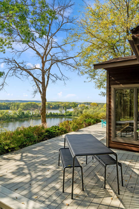 Outdoor deck with a river view, featuring a metal table and chairs, framed by trees and greenery.