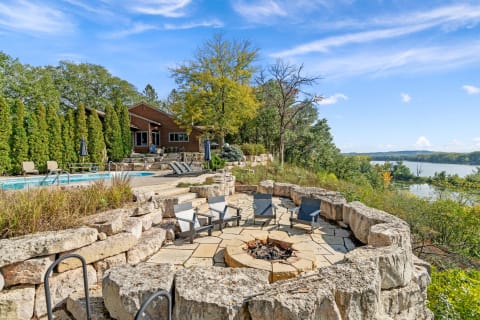 Outdoor landscape with a pool, fire pit, and a wooden house set against a river view.