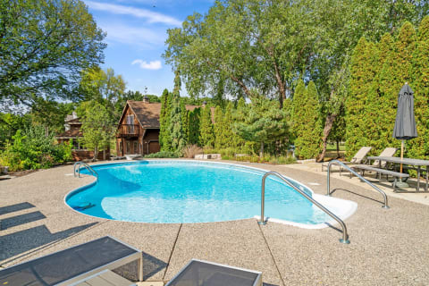 Outdoor swimming pool with turquoise water, concrete surroundings, and wooden house in the background.