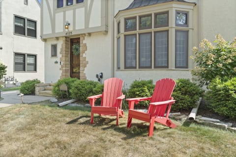 Two red Adirondack chairs in a sunny front yard beside a Tudor-style home with lush landscaping.