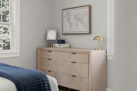 A soft gray bedroom corner with a wooden dresser, brass lamp, circular mirror, and vintage map on the wall.