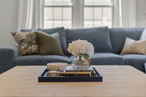 A cozy living room featuring a coffee table with a black tray, a glass vase of white flowers, books, and a plush blue couch with decorative pillows.