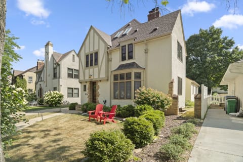 Tudor-style house with landscaped front yard featuring red chairs and flowering bushes.