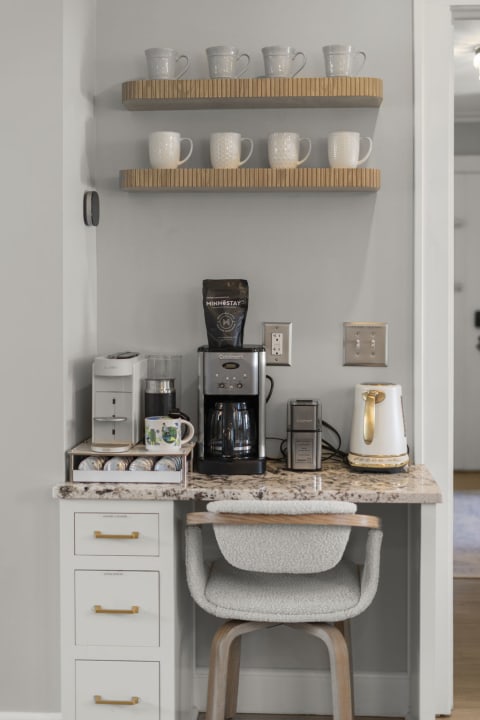 A modern coffee bar featuring a coffee maker, kettle, mugs on wooden shelves, and a stylish chair.