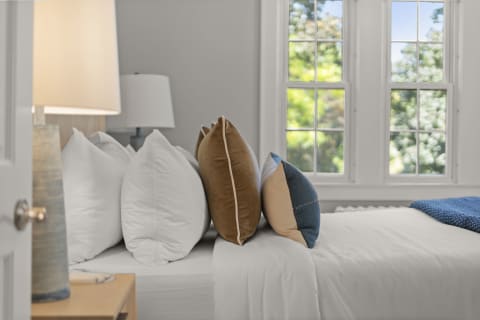 A well-decorated bedroom featuring white linens, colorful pillows, and soft natural light from the windows.