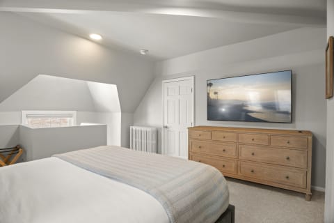 A cozy bedroom with vaulted ceilings, a textured quilt on the bed, wooden dresser, and a beach scene on the TV.