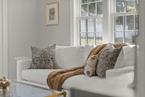 A white sofa with decorative pillows and a chunky knit blanket, illuminated by sunlight from large windows.