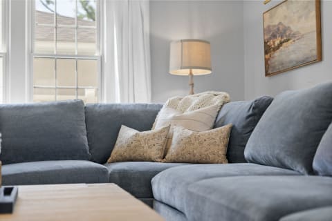 A cozy living room corner featuring a blue sectional couch with decorative pillows, a knit throw, a lamp, and a scenic painting on the wall.