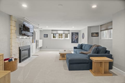 A spacious lower-level living room with a plush blue sectional sofa, stone fireplace, and mounted television.