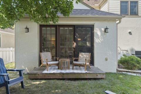 A cozy outdoor deck with wooden chairs, a round table, and a striped rug, surrounded by trees and bright light.