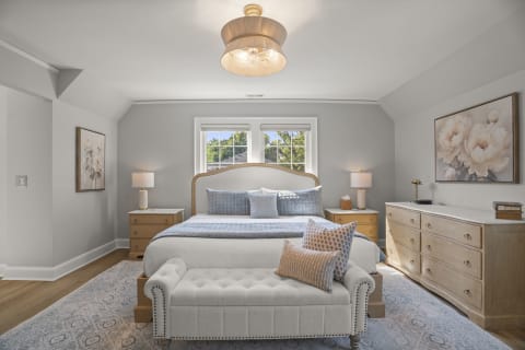 An elegant bedroom featuring a plush bed with soft linens, warm wood furniture, and abundant natural light.