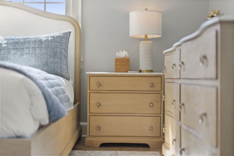 A cozy bedroom featuring a light wood bed, a quilted blue throw, a stylish nightstand with a lamp, and soft natural lighting.
