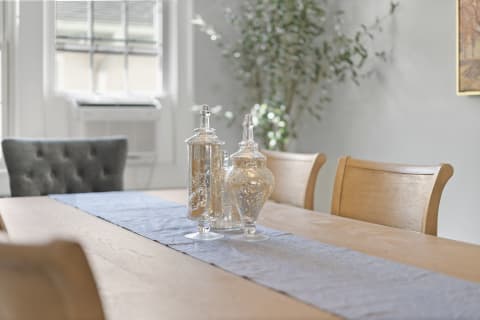 A dining table with a blue runner and decorative glass jars, illuminated by natural light, featuring a tufted gray chair and greenery in the background.