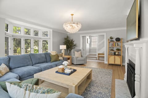 Modern living room with blue sectional, wooden coffee table, and elegant chandelier, featuring natural light from large windows.