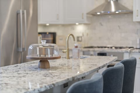Modern kitchen with a granite island, featuring a glass cake dome and elegant bar stools.