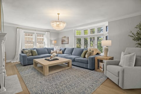 A spacious living room featuring blue sofas, a chandelier, and large windows with greenery outside.