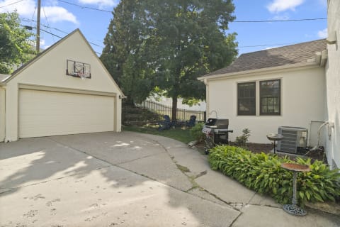 A private driveway next to a garage, featuring a grill and basketball hoop, surrounded by greenery.
