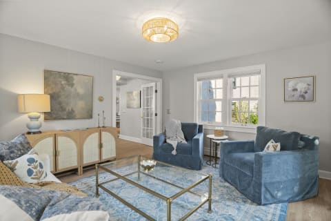 A cozy living room with plush blue chairs, glass table, and soft lighting, brightened by sunlight through French doors.