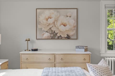 A serene bedroom showcasing a dresser with a floral painting, soft decor, and natural light coming through a window.