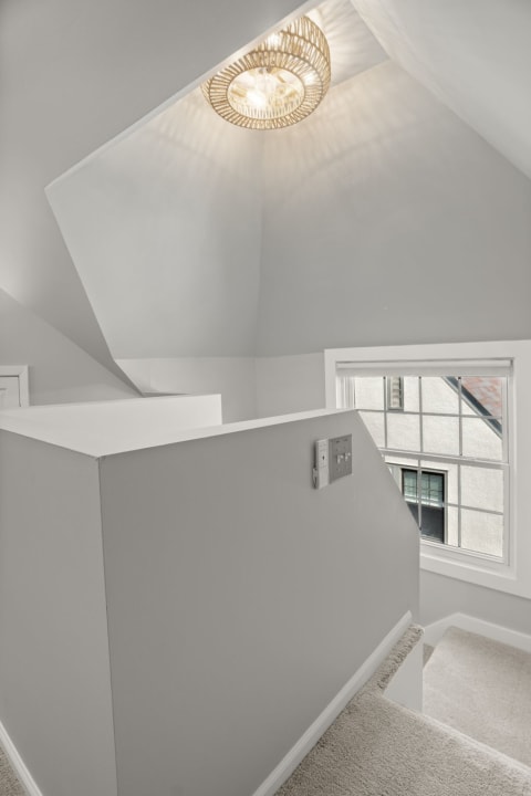 A bright, airy hallway featuring a vaulted ceiling and woven pendant light, with a window showing soft daylight.