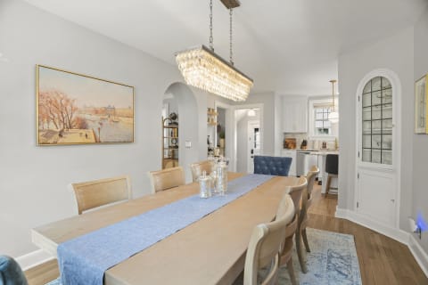 Elegant dining area featuring a long wooden table, crystal chandelier, and wall art depicting a scenic waterfront.