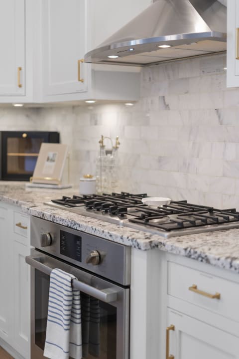 Modern kitchen featuring a gas range with stainless steel hood, marble backsplash, and white cabinetry.
