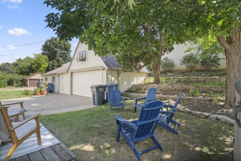 A peaceful outdoor space featuring blue Adirondack chairs, a stone wall garden, and a garage with an outdoor basketball hoop.