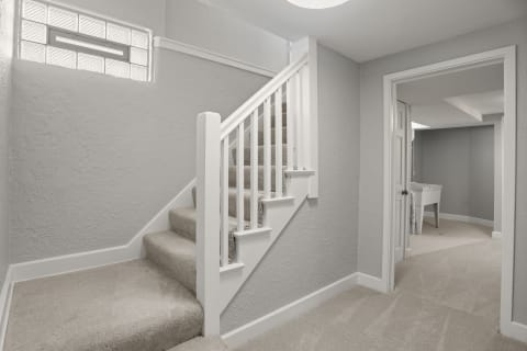 A light gray hallway featuring a staircase with white railings and beige carpet, illuminated by natural light.