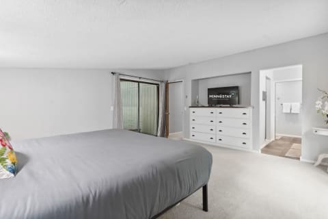 A spacious bedroom featuring a gray bedspread, a colorful pillow, a white dresser with a TV, and natural light coming from a sliding door.