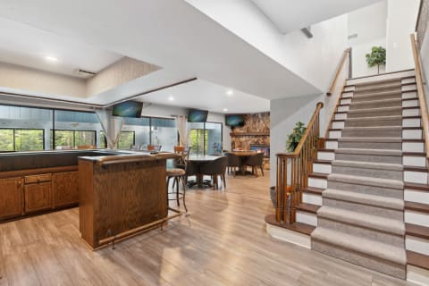 A spacious modern interior with a bar, dining area, and fireplace, featuring large windows and a staircase.