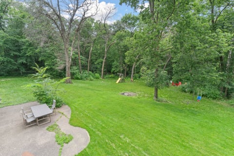 Backyard view featuring grass, a swing set with a slide, seating area, and dense trees.