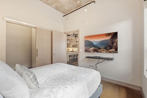 Modern bedroom with a bed, mounted TV showing mountain landscape, and doorway to kitchen.