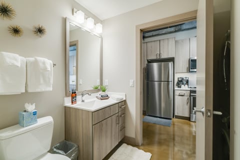 Stylish bathroom with white towels, large mirror, and light wood vanity connected to a kitchen.