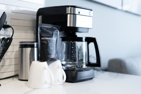 Sleek black and stainless steel coffee maker beside two white mugs and a coffee bag on a marble countertop.